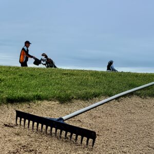 Two golfers walking on a course with a rake in a sand bunker under a cloudy sky.